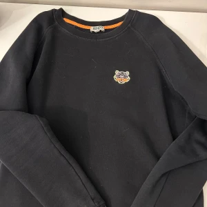 Svart sweatshirt från Kenzo med tiger - Svart sweatshirt från Kenzo i bomull med klassisk rund hals och broderad tigerlogga på bröstet. Snygg orange detalj på insidan av kragen. Perfekt för dig som gillar streetstyle och vill ha något som sticker ut.