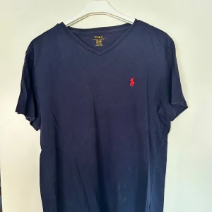 Mörkblå t-shirt Polo Ralph Lauren - Snygg mörkblå t-shirt från Polo Ralph Lauren med klassisk röd logga broderad på bröstet. T-shirten har v-ringning och korta ärmar, tillverkad i mjuk bomull. Perfekt för en clean och stilren look. Nypris 699kr