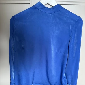 g blå blus i stl 38 från NAKD, volangärm & hög hals - Superfin blus i storlek 38 från Andrea Hedenstedts kollektion för NA-KD. Långärmad modell med eleganta volangärmar och stilren hög hals. Knappar i nacken som fin detalj i ryggen. Perfekt både till vardags och fest!