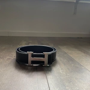 Svart skärp med silvrigt H-spänne - Snyggt svart skärp från med ikoniskt H-spänne i silverfärgad metall. Bältet har vita kontrastsömmar längs kanterna och är gjort i äkta skinn. Perfekt accessoar för att lyfta vilken outfit som helst.