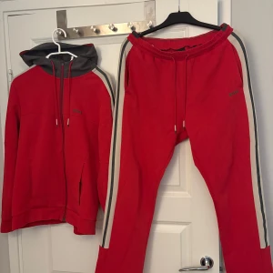 Röd Hugo Boss dress - Röda mjukisbyxor från BOSS i strl M med grå och vita revärer längs sidorna. Byxorna har elastisk midja med snörning och är tillverkade i mjuk bomull. Perfekta för chill eller träning, med en clean och sportig vibe. Säljs tillsammans med en matchande zip hoodie som är i strl xs/kan sitta okej på nån med S. Inga deflekter eller liknande saker. Tröjan går inte att köpa i butik. Därav priset. Priset går att diskuteras och kan köpas enskilt.
