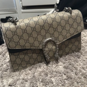 Gucci Dionysus axelväska med kedja - Säljer en Gucci Dionysus axelväska i beige med klassiskt GG-mönster. Väskan har en snygg silverfärgad kedja och ett detaljerat spänne framtill. Insidan är röd med dragkedjefack