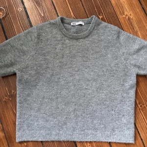 Grå stickad tröja från Zara - Mysig grå stickad tröja från Zara med rund halsringning och ribbade muddar i storlek XL men känns som en M. Tröjan har kortare ärmar och en rak, lite boxig passform som ger en chill vibe. Perfekt att styla med jeans eller kjol för en enkel men snygg look.