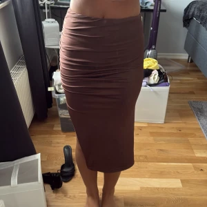 Brun tight långkjol med slits - Säljer en brun långkjol i tight passform med snygg rynkning på ena sidan och en hög slits. Kjolen är figurnära och har en mjuk, stretchig känsla som sitter skönt på kroppen. Perfekt för dig som gillar en stilren look med lite edge.