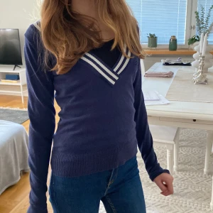 Mörkblå v-ringad stickad tröja - Mörkblå stickad tröja med v-ringning och vita ränder runt halsen. Tröjan har lång ärm, ribbade muddar och ett broderat emblem på bröstet för en preppy look. Perfekt till jeans för en chill och snygg stil.