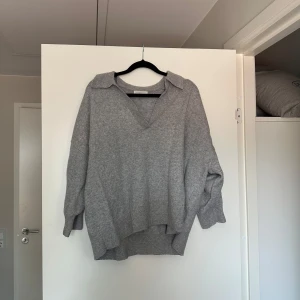 Grå stickad v-ringad tröja H&M - Mysig grå stickad tröja från H&M med v-ringning och krage. Tröjan har en loose passform och långa ärmar, perfekt för lager på lager. Ribbstickade muddar och lite längre baktill för en avslappnad look.