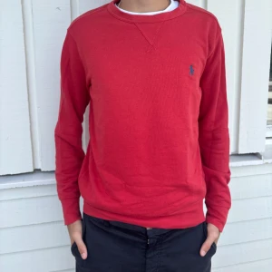 Polo Ralph Lauren tröja - Röd långärmad tröja från Polo Ralph Lauren med klassisk rund halsringning och blå broderad logga på bröstet. Tillverkad i en mjuk mix av bomull, polyester och modal. Perfekt för dig som gillar stilren och enkel design.