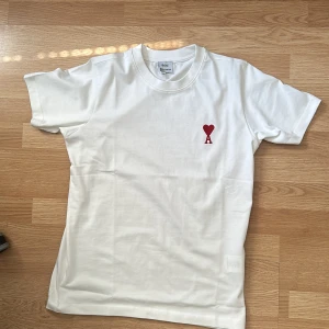 Vit t-shirt från Ami Paris med hjärtdetalj - Vit t-shirt från Ami Paris i mjuk bomull. Klassisk passform med korta ärmar och rund halsringning. På bröstet finns en röd broderad hjärtdetalj med bokstaven A. Perfekt för en clean och stilren look. Säljer den eftersom den är för stor. Storlek S