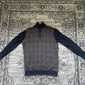 Mörkblå stickad tröja Stefano Ricci - Exklusiv mörkblå stickad tröja från Stefano Ricci med halv dragkedja och hög krage. Framsidan har ett snyggt geometriskt mönster i blått och grått. Långa ärmar och diskret metallogo vid midjan. Perfekt för dig som gillar stilren och lyxig design.