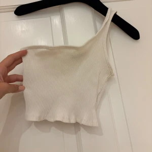 Vit ribbad oneshoulder topp - Snygg vit ribbad topp med oneshoulder-design. Croppad modell som sitter tight och har en cool asymmetrisk axel. Perfekt till jeans eller kjol för en trendig look.