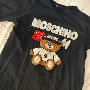 Svart Moschino t-shirt med nalle - Svart t-shirt från Moschino med stort broderat nallemotiv och logga framtill. T-shirten har rund hals, korta ärmar och coola 3D-detaljer i text och applikation. Perfekt statement-plagg för dig som gillar streetstyle och lekfull design. Endast använd några gånger, storlek medium