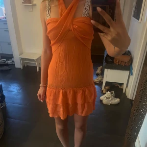 Orange kort klänning med volang - Supersöt orange kortklänning med omlott-detalj upptill och volangkant nertill. Klänningen har axelband med virkade detaljer som man knyter runt halsen och en snygg, somrig look. Perfekt för varma dagar och ger en riktigt fräsch vibe.