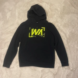 Svart hoodie från Warp  - Svart hoodie från Warp med stor huva och lång ärm. Framtill finns ett neongult tryck med grafiska detaljer och Warp-logga. Tröjan är i mjuk bomullsblandning och har en klassisk, avslappnad passform. 