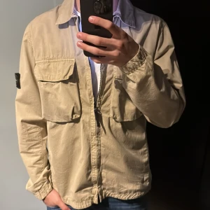 Beige overshirt från Stone Island - Snygg beige overshirt från Stone Island med klassisk patch på ärmen. Jackan har två stora bröstfickor, dragkedja framtill och avslappnad passform. Hör av dig med frågor!