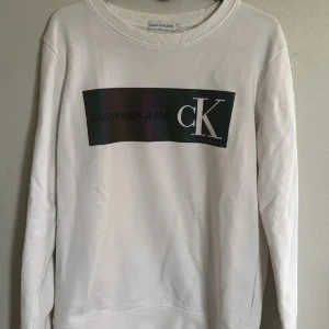 Vit Calvin Klein Jeans sweatshirt - Vit sweatshirt från Calvin Klein Jeans med svart logga och CK-tryck på bröstet. Klassisk rund halsringning och långa ärmar, perfekt för en clean och stilren look.