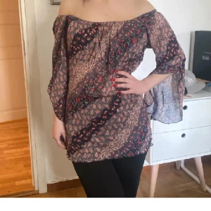 Blommig mesh offshoulder blus med slitsärm - Säljer en flowy blus med offshoulder och vida slitsärmar. Toppens mönster är en mix av små blommor och leopard i bruna, svarta och röda toner. Tunt, transparent material som ger en luftig känsla. Perfekt för dig som gillar boho-vibe och unika detaljer. Tryck ej köp nu