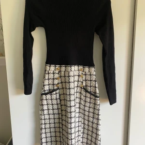 Svart och vit klänning från Lipsy London - Supersnygg klänning från Lipsy London med ribbad svart överdel och långärmade ärmar. Kjoldelen är vit med svart rutigt mönster, dekorerad med guldknappar och svarta fickdetaljer. OBS!!!!!Dragkedjan kan krångla lite ibland 