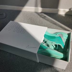 Turkosa sneakers från Nike By You - Unika turkosa sneakers från Nike By You med snygga detaljer i silver och vit sula. Skorna har snörning och är tillverkade i syntetmaterial med mesh för extra andningsförmåga. Perfekta för dig som vill sticka ut med en personlig stil.