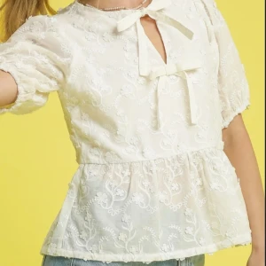 Vit blommig blus med rosetter - Supersöt vit blus med broderade blommor och två rosetter framtill. Blusen har korta puffärmar och volang nedtill för en romantisk vibe. Perfekt att styla med jeans eller kjol för en trendig look.