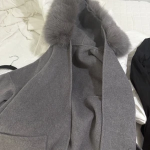 Grå kappa med rävpäls XS - Snygg grå kappa i ullmix med stor huva kantad av fluffig grå rävpäls. Jackan har raka ärmar och en enkel, stilren design utan synliga knappar eller dragkedja. Perfekt för dig som gillar en clean look med lite extra lyxig känsla. Köpt för ca 4000, använd fåtalgånger inga synliga skador.
