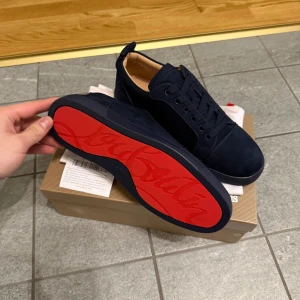 Mörkblå sneakers från Christian Louboutin - Säljer ett par mörkblå sneakers från Christian Louboutin med klassisk röd sula och snörning. Skorna är i mocka och har en rund tå samt platt sula. Perfekta för dig som vill ha något stilrent men ändå sticker ut med den ikoniska röda sulan.