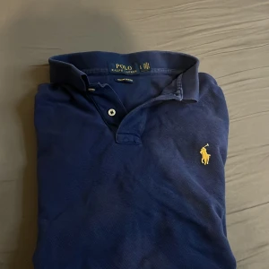 Marinblå pikétröja från Ralph Lauren - Klassisk marinblå pikétröja från Ralph Lauren med gul broderad logga på bröstet. Tröjan har krage, två knappar och korta ärmar. Tillverkad i mjuk bomull som är skön mot huden. Perfekt för en stilren och avslappnad look. Hör av er vid minsta lilla fråga 