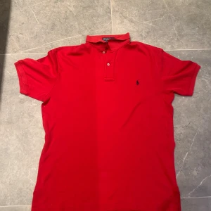 Röd pikétröja från Polo Ralph Lauren - Storlek s men skulle säga att den sitter mer som M Klassisk röd pikétröja från Polo Ralph Lauren i 100% bomull. Tröjan har korta ärmar, krage och två knappar framtill. Den ikoniska broderade loggan i mörkblått syns på bröstet. Perfekt för en stilren och sportig look.