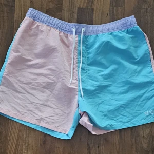 Pastellblå och rosa badshorts - Never Compromise - Snygga badshorts i pastellblått och ljusrosa från Never Compromise. De har elastisk midja med vit snörning, bakficka med lock och broderad logga. Perfekta för stranden eller poolen, med en loose fit och lätt syntetmaterial som torkar snabbt.