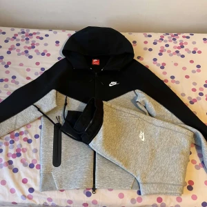 Nike hoodie jacka svart/grå - Nike hoodie jacka i svart och ljusgrått med dragkedja hela vägen och två stora fickor med svarta detaljer. Jackan har huva med snörning och klassisk Nike-logga på bröstet. Perfekt för en sportig och avslappnad stil.