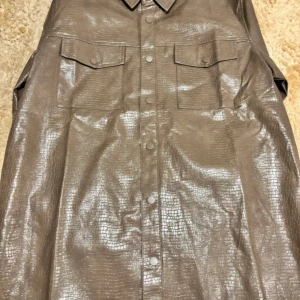 Brun overshirt i skinnlook  - Cool brun overshirt från Reclaimed Vintage med croc-mönstrad yta i skinnlook. Skjortan har två bröstfickor med lock, tryckknappar framtill och klassisk krage. Materialet är polyuretan på utsidan och viskos på insidan. Perfekt för dig som vill sticka ut. Endast testad!! Säljs då vi inte hann skicka tillbaks för att byta färg. Nypris är 400.