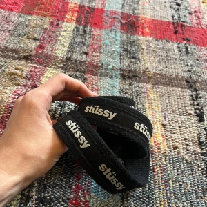 Svart Stüssy tygbälte med logga - Svart tygbälte från Stüssy med vit logga tryckt längs hela bältet. Bältet har en klassisk D-ring i metall som spänne och är gjort i slitstarkt material. Perfekt accessoar för att lyfta streetwear-looken.