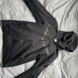 Nike tech  - Svart hoodiejacka från Nike med hel dragkedja och huva. Jackan har diskreta svarta detaljer över bröstet och en praktisk ficka med dragkedja på ena ärmen. Perfekt för dig som gillar en clean och sportig stil.