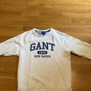 Vit sweatshirt från GANT, storlek M - Vit sweatshirt från GANT med blå text 'GANT 1949 NEW HAVEN' tryckt på bröstet. Klassisk rund halsringning och långa ärmar. Mjuk insida och ribbade muddar. Perfekt för en clean och clasy look.