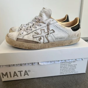 Vita sneakers från Premiata, strl 41 - Snygga vita sneakers från Premiata med grå snörning och svart detalj på hälen. Skorna har en rund tå, platt sula och är tillverkade i skinn. Klassisk design med logga på sidan och coola detaljer på sulan. Perfekta för dig som gillar streetstyle.