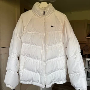 Vit pufferjacka från Nike - Säljers en riktigt snygg och tidlös vit pufferjacka från Nike med diskret svart logga på bröstet och ryggen. Jackan har hög krage, dragkedja framtill och breda, quiltade partier som ger en cool och sportig vibe. Perfekt för kalla dagar när du vill hålla stilen. Ej tvättat som syns på bild 3 men går säkert bort i tvätt eller klorin. Oversize så likt en L