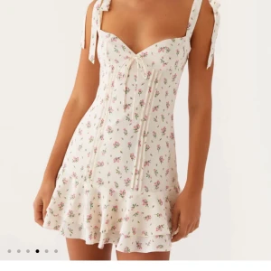 Vit blommig kortklänning med volang - Supersöt vit kortklänning med små rosa blommor, volangkant och knytband på axlarna. Klänningen har v-ringning och en feminin passform som framhäver midjan. Perfekt för varma dagar och somriga vibes!