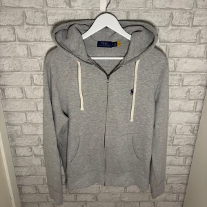 Ralph Lauren zip - Ralph Lauren zip med dragkedja, huva och vita snören. Tröjan har broderad blå logga på bröstet och kängurufickor framtill. Tillverkad i mjuk bomullsblandning för en skön och avslappnad stil. Använt skick, lite knopprig. Hör av dig vid intresse! 🙌