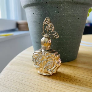 Anna Sui tom parfymflaska med rosor - Superfin tom parfymflaska från Anna Sui i guldig metall-look, formad som två rosor med ett detaljerat fjärilsmönster på korken. Flaskan är kompakt och har en lyxig, romantisk design som verkligen sticker ut i samlingen.