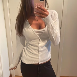 Vit kofta med v-ringning och knappar - Snygg vit kofta med v-ringning och knappar framtill. Modellen är figurnära med långa ärmar och ribbade muddar vid ärmslut och nederkant. Perfekt att slänga över ett linne eller topp för en clean och fräsch look.