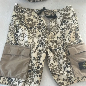 Stone Island alligator camo cargo shorts limited edition 2018 - Snygga camouflage-mönstrade cargo shorts från Stone Island i beige och bruna toner. Shortsen har stora fickor på sidorna, snörning i midjan och klassisk Stone Island patch på ena benet. Tillverkade i polyester, perfekta för sommaren och streetwear-stil.                                       Kan köpas tillsammans med t-shirten som tillhör i ett set till ett bättre pris.