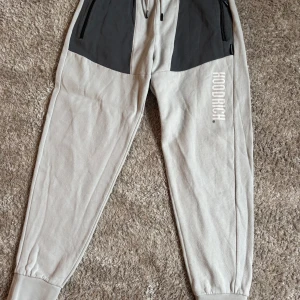 Grå mjukisbyxor från Hoodrich L - Snygga grå mjukisbyxor från Hoodrich i storlek L. Byxorna har svarta detaljer upptill, dragsko i midjan och muddar vid bensluten. Stort Hoodrich-tryck på ena benet och bakficka med logga. Perfekta för chill eller streetwear.