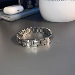 Silverfärgat armband i metall - Stilrent armband i silverfärgad metall med rektangulära länkar och graverade detaljer. Armbandet har ett diskret mönster på vissa länkar och en ingraverad fjäder på en av plattorna. Perfekt accessoar för en clean och modern look.