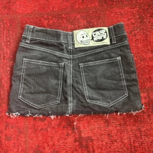 Svart lågmidjad jeanskjol från Cheap Monday - Svart lågmidjad jeanskjol från Cheap Monday med rå, fransig kant och tydliga vita sömmar. Klassisk femficksmodell med stor logopatch bak. Gjord i bomull, polyester och lite stretch för skön passform. Perfekt till sommaren. Tryck ej köp nu