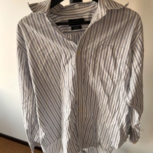 Randig skjorta från Ralph Lauren - Klassisk vit skjorta med blå ränder från Ralph Lauren. Skjortan har lång ärm, bröstficka och knappar framtill. Tillverkad i mjuk bomull och har en avslappnad passform. Perfekt för en stilren och tidlös look.