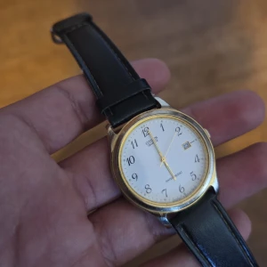 Citizen klassisk armbandsklocka guld/vit - Citizen armbandsklocka med vit urtavla, svarta siffror och datumvisning. Boetten är i guldtonad metall och bandet är svart, gjort av läder. Stilren och tidlös design med tunna visare i guld. Perfekt för dig som gillar klassiska accessoarer.