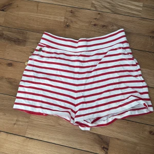 Randiga shorts från Holly & Whyte - Snygga högmidjade shorts från Holly & Whyte med röda och vita horisontella ränder. De har dekorativa knappar i sidan och fickor bak. 