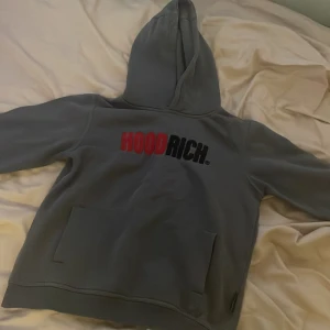 Grå HOODRICH hoodie med tryck - Snygg grå hoodie från HOODRICH med stor ficka framtill och huva. Framsidan har ett tydligt tryck med texten HOODRICH i rött och svart. Perfekt för dig som gillar streetwear och vill ha en bekväm och stilren look.