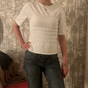 Vit smockad topp med kort ärm NELLY - Säljer en vit topp med smockad effekt över hela magen och ryggen. T-shirten har rund halsringning och korta ärmar, sitter figurnära och är superenkel att matcha till jeans eller kjol. Perfekt för dig som gillar stilrena och bekväma plagg. XS men känns som S