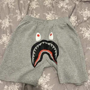 Grå shorts med hajmotiv - Grå mjuka shorts med elastisk midja och coolt hajtryck framtill. Shortsen har fickor på sidorna och är tillverkade i ett bekvämt bomullsmaterial. Perfekta för chill dagar eller när du vill sticka ut lite extra.