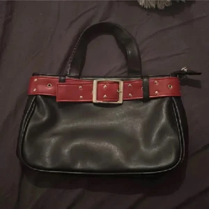 VINTAGE❤️Svart handväska med röd bältdetalj - Cool svart handväska i skinnimitation med röd bältdetalj och stora guldfärgade nitar. Väskan har två handtag och stängs med dragkedja upptill. Perfekt för dig som vill sticka ut med en edgy accessoar.